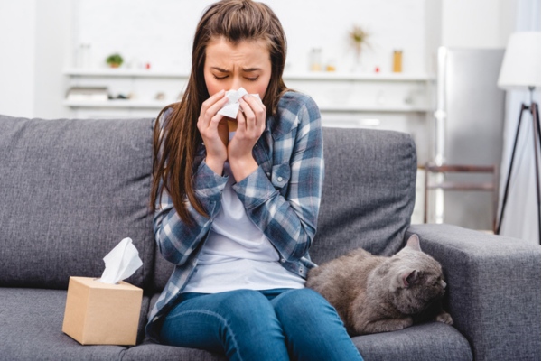 woman blowing her nose on the couch beside a cat depicting poor indoor air quality