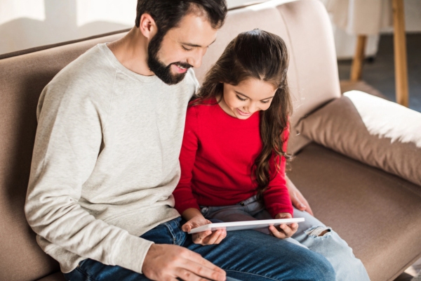 Father and daughter sitting on sofa while looking at smart tablet and feeling warm and cozy