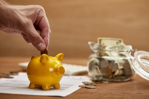 Cropped view of man's hand putting coin in a golden piggy bank with money in a jar in the background depicting Reduced Energy Expenses