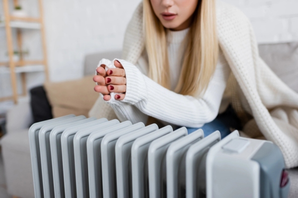 Cropped view of young blonde woman covered in blanket keeping warm on an electric heater due to running out of heating oil