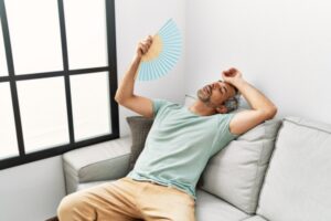 man using hand fan while sitting on the couch due to air conditioning not working properly