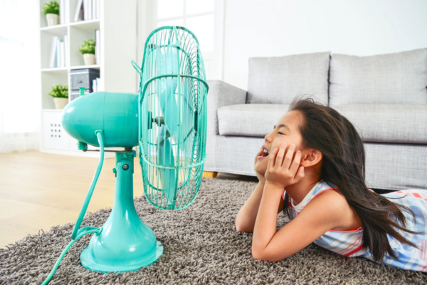 kid lying on a carpeted floor in front of a green fan due to AC not keeping up